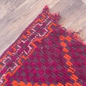 Pode incluir: Close-up de um tapete vibrante e texturizado com um padr&atilde;o geom&eacute;trico. O tapete apresenta uma rica paleta de cores de magenta profundo, laranja e tons de rosa. As bordas s&atilde;o franjadas, adicionando um toque de textura e detalhe ao design.