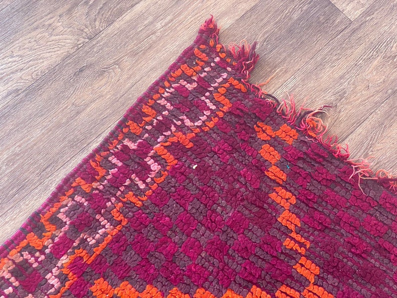 Pode incluir: Close-up de um tapete vibrante e texturizado com um padr&atilde;o geom&eacute;trico. O tapete apresenta uma rica paleta de cores de magenta profundo, laranja e tons de rosa. As bordas s&atilde;o franjadas, adicionando um toque de textura e detalhe ao design.