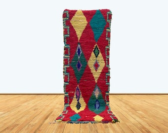 May include: A vibrant, rectangular rug with a red background and diamond patterns in yellow, teal, and blue. The rug has a geometric border and is displayed on a wooden floor.