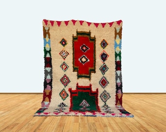 May include: A beige, rectangular rug with a central red and green geometric design. The rug features colorful diamond patterns and a border with various colors, including blue, brown, and pink. The rug is displayed on a wooden floor.