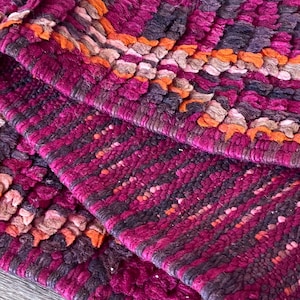Pode incluir: Close-up de um tapete texturizado com uma borda dobrada. O tapete apresenta um padr&atilde;o de tons roxo escuro, magenta, laranja e marrom. O design tecido cria uma superf&iacute;cie t&aacute;til, adequada para decora&ccedil;&atilde;o de interiores.
