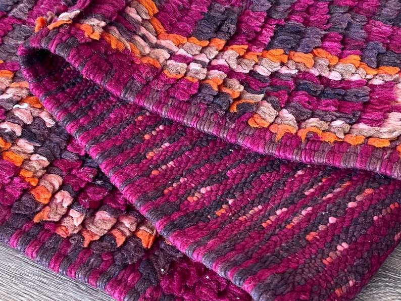Pode incluir: Close-up de um tapete texturizado com uma borda dobrada. O tapete apresenta um padr&atilde;o de tons roxo escuro, magenta, laranja e marrom. O design tecido cria uma superf&iacute;cie t&aacute;til, adequada para decora&ccedil;&atilde;o de interiores.
