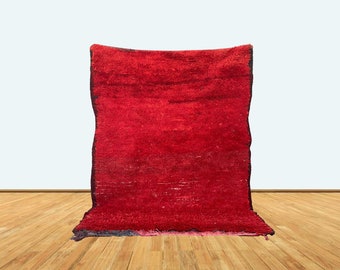 May include: A rectangular area rug featuring a deep, solid red color with a thick, shaggy pile texture. The rug has dark, visible edges along the sides and a slightly uneven, hand-woven appearance throughout the surface of the fabric.