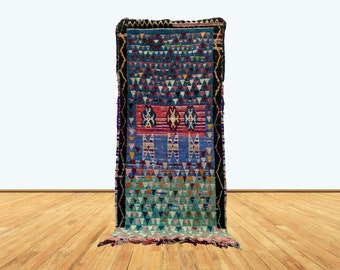 Puede incluir: Una alfombra rectangular tejida a mano con un diseño geométrico vibrante. La alfombra presenta una paleta de colores de azul, rojo, naranja y verde, con un borde negro. El patrón incluye triángulos y rombos.