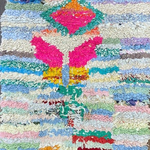 Puede incluir: Alfombra de trapo hecha a mano con un dise&ntilde;o geom&eacute;trico. La alfombra presenta una vibrante mezcla de tiras de tela rosas, verdes, azules y crema. El dise&ntilde;o incluye una forma de diamante central y patrones sim&eacute;tricos.