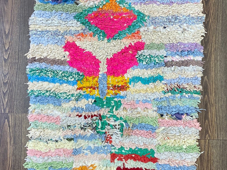 Puede incluir: Alfombra de trapo hecha a mano con un dise&ntilde;o geom&eacute;trico. La alfombra presenta una vibrante mezcla de tiras de tela rosas, verdes, azules y crema. El dise&ntilde;o incluye una forma de diamante central y patrones sim&eacute;tricos.