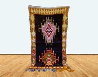 May include: A colorful Moroccan rug with a dark blue background and intricate geometric patterns in shades of red, yellow, purple, and white. The rug has a fringe border and is displayed on a wooden floor.