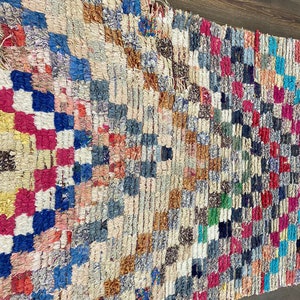 May include: A colorful Moroccan rug with a geometric pattern in shades of blue, red, pink, yellow, and brown. The rug is made of wool and has a hand-woven texture.