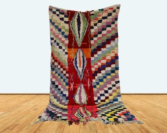 May include: A colorful Moroccan rug with a geometric pattern and a central design of stylized teardrop shapes. The rug is woven with wool and has a fringe on the edges.