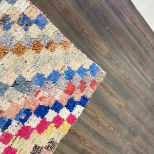 May include: A colorful, handwoven rug with a geometric pattern in shades of blue, pink, yellow, and white. The rug is laid on a dark brown wood floor.