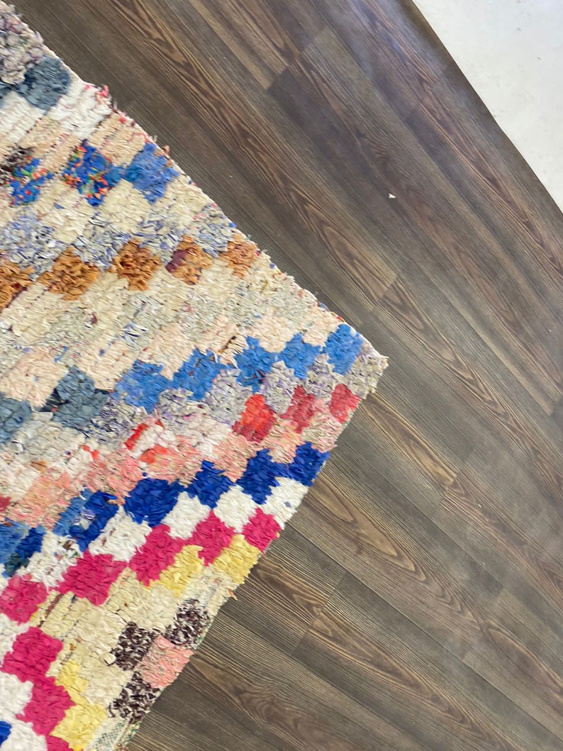 May include: A colorful, handwoven rug with a geometric pattern in shades of blue, pink, yellow, and white. The rug is laid on a dark brown wood floor.