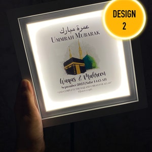 May include: A white framed illuminated sign with a watercolor illustration of the Kaaba, a mosque in Mecca, Saudi Arabia. The sign reads "Ummrah Mubarak" in Arabic and "Waqas & Mahreen September 2023 Safar 1445 AH "And complete the Hajj and Umrah for Allah" (Quran 2:196).