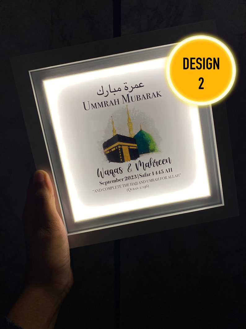 May include: A white framed illuminated sign with a watercolor illustration of the Kaaba, a mosque in Mecca, Saudi Arabia. The sign reads "Ummrah Mubarak" in Arabic and "Waqas & Mahreen September 2023 Safar 1445 AH "And complete the Hajj and Umrah for Allah" (Quran 2:196).