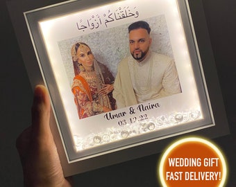 May include: A white light-up picture frame with a photo of a couple in their wedding attire. The frame has a white border and a clear plastic front. The photo shows the couple smiling and looking at each other. The frame also has a small inscription that reads "Umar & Naira 03.12.22".