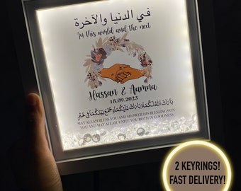 May include: A white light-up frame with a floral design and the text "In this world and the next" in English and Arabic. The names "Hassan & Aamna" and the date "18.09.2023" are also included. The frame is decorated with small, clear rhinestones.