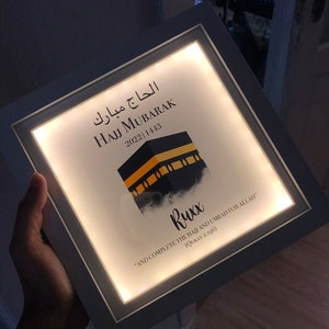 May include: A white framed light-up sign with the text "Hajj Mubarak 2022 1443" in Arabic and English. The sign also features a stylized image of the Kaaba, a cube-shaped building in Mecca, Saudi Arabia, and a quote in English: "And complete the Hajj and Umrah for Allah" (Quran 2:196).