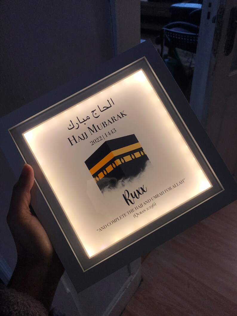 May include: A white framed light-up sign with the text "Hajj Mubarak 2022 1443" in Arabic and English. The sign also features a stylized image of the Kaaba, a cube-shaped building in Mecca, Saudi Arabia, and a quote in English: "And complete the Hajj and Umrah for Allah" (Quran 2:196).
