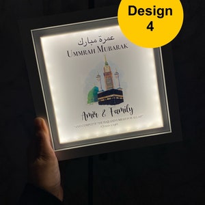 May include: A white framed illuminated sign with a watercolor illustration of the Kaaba in Mecca. The sign reads "Ummrah Mubarak" in Arabic and English, and "Amir & Family" with a Quran verse about completing the Hajj and Umrah.