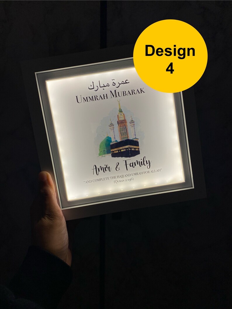 May include: A white framed illuminated sign with a watercolor illustration of the Kaaba in Mecca. The sign reads "Ummrah Mubarak" in Arabic and English, and "Amir & Family" with a Quran verse about completing the Hajj and Umrah.