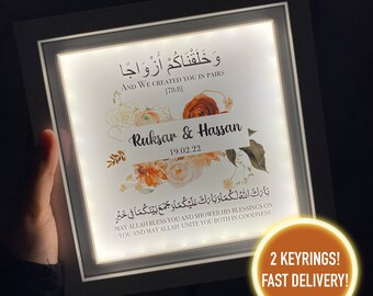 May include: A white framed illuminated sign with a floral design and the Arabic text "وَخَلَقْنَاكُمْ أَزْوَاجًا" which translates to "And We Created You in Pairs [78:8]"  The sign also includes the names "Ruksar & Hassan" and the date "19.02.22".  The text "بَارَكَ اللهُ لَكُمَا وَبَارَكَ عَلَيْكُمَا وَ جَمَعَ بَيْنَكُمَا فِي خَيْرٍ" which translates to "May Allah bless you and shower His blessings on you and may Allah unite you both in goodness" is also included.  The sign has a gold circle with the text "2 KEYRINGS! FAST DELIVERY!"