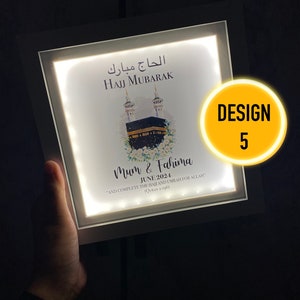 May include: A white light-up frame with a picture of the Kaaba, a mosque in Mecca, Saudi Arabia. The frame has the text "Hajj Mubarak" in Arabic and English, and the names "Mum & Fahima" with the date "June 2024". The text "And complete the Hajj and Umrah for Allah (Quran 2:196)" is also included.