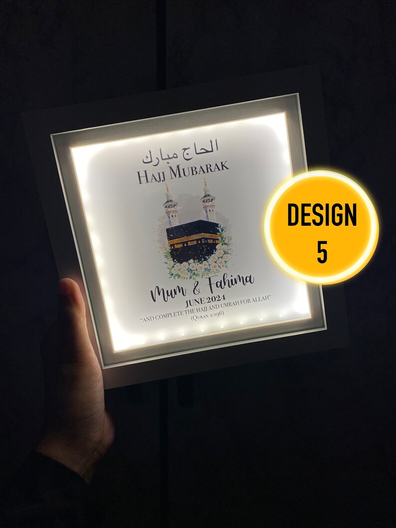May include: A white light-up frame with a picture of the Kaaba, a mosque in Mecca, Saudi Arabia. The frame has the text "Hajj Mubarak" in Arabic and English, and the names "Mum & Fahima" with the date "June 2024". The text "And complete the Hajj and Umrah for Allah (Quran 2:196)" is also included.