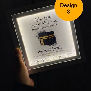 May include: A white framed illuminated picture with a white background. The picture features a watercolor painting of the Kaaba, a mosque in Mecca, Saudi Arabia. The text "Umrah Mubarak" is written in Arabic and English. The text "May 2023 | Shawwal 1444 AH" is written below the Arabic text. The text "Mahmood Family" is written below the image. The text "And complete the Hajj and Umrah for Allah" is written below the family name. The text "(Quran 2:196)" is written below the quote. The picture is decorated with small, clear rhinestones.