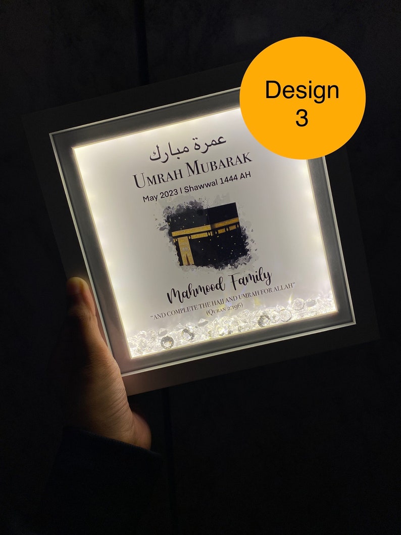 May include: A white framed illuminated picture with a white background. The picture features a watercolor painting of the Kaaba, a mosque in Mecca, Saudi Arabia. The text "Umrah Mubarak" is written in Arabic and English. The text "May 2023 | Shawwal 1444 AH" is written below the Arabic text. The text "Mahmood Family" is written below the image. The text "And complete the Hajj and Umrah for Allah" is written below the family name. The text "(Quran 2:196)" is written below the quote. The picture is decorated with small, clear rhinestones.