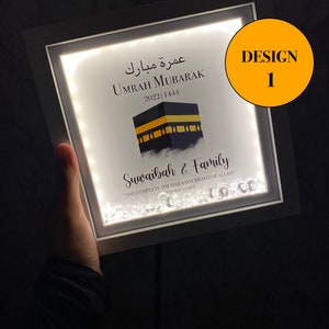 May include: A white framed light-up sign with a black and gold image of the Kaaba, the Islamic holy site in Mecca, Saudi Arabia. The sign reads "Umrah Mubarak 2022 1444" in Arabic and English. Below the image is the text "Suwaibah & Family" and a quote from the Quran: "And complete the Hajj and Umrah for Allah" (Quran 2:196). The sign is decorated with small, clear crystals.