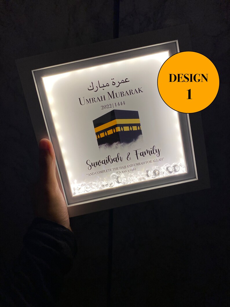 May include: A white framed light-up sign with a black and gold image of the Kaaba, the Islamic holy site in Mecca, Saudi Arabia. The sign reads "Umrah Mubarak 2022 1444" in Arabic and English. Below the image is the text "Suwaibah & Family" and a quote from the Quran: "And complete the Hajj and Umrah for Allah" (Quran 2:196). The sign is decorated with small, clear crystals.