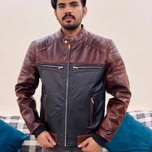 Puede incluir: Chaqueta de cuero de dos tonos, marrón en la parte superior y negra en la inferior. La chaqueta tiene cuello alzado, bolsillos con cremallera y cierre frontal con cremallera. Las mangas son marrones con detalles acolchados.