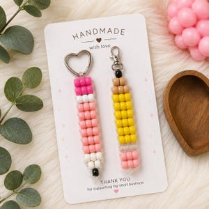May include: Two pencil-shaped keychains made of colorful plastic beads attached to a white display card with the text HANDMADE with love and THANK YOU for supporting my small business. One keychain has pink and white beads, the other yellow and tan.