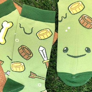 May include: A pair of light green socks featuring a pixelated pattern of yellow bones, brown barrels, gold coins, white swords, and arrows. One sock displays a simple, smiling face with two black eyes and a curved mouth near the dark green toe.