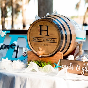 May include: A wooden barrel with a black and white striped design, personalized with the initials "H" and the names "Jackson & Daniella" and the date "6.14.20". The barrel is on a wooden stand and has a spigot. There is a sign that says "cards & gifts" and a blue and white wooden sign with the letters "J" and "D" and the word "Love" in between.