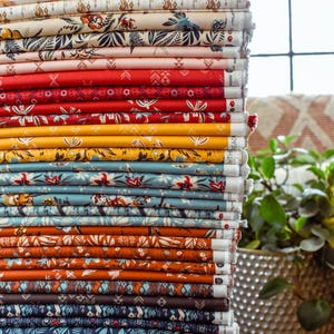 May include: A tall stack of folded fabric bolts featuring a diverse array of patterns including floral, geometric, and botanical prints in a vibrant color palette ranging from cream and soft pink to deep red, mustard yellow, teal, and dark navy blue.