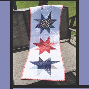 May include: A table runner with a white background and three red, white, and blue patchwork stars. The text "Crazy Stars" is at the top of the image. The text "Tricia Lynn Maloney Orphan Quilt Designs For Villa Rosa Designs" is at the bottom of the image.