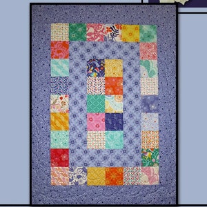 May include: A colorful quilt with the word "DAYBREAK" in black letters. The quilt features a central rectangle of patterned squares in various colors, surrounded by a blue floral border. The words "Villa Rosa Designs" are at the bottom.