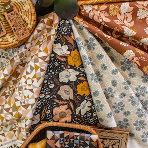 Puede incluir: Surtido de muestras de tela estampadas en varios colores, incluyendo marrón, amarillo y negro, con diseños florales y geométricos. Una cesta de mimbre contiene tela adicional y una caja de madera. También se ve un llavero de cuero con el texto "Amazing Grace".