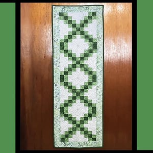 May include: A green and white table runner with a shamrock design. The table runner is called "Erin" and is an orphan quilt design by Tricia Lynn Maloney for Villa Rosa Designs.