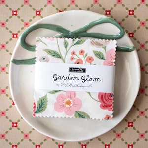 Puede incluir: Una pila de muestras de tela cuadradas con bordes dentados que presentan un patrón floral de flores rosas y coral con hojas verdes. Una banda de papel blanco envuelve el centro con el texto Garden Glam by Lella Boutique y Moda.