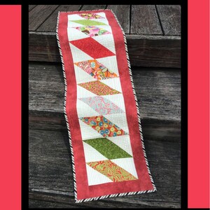 May include: A red, white, and green diamond-shaped table runner with a black and white trim. The table runner is on a wooden surface. The text "Diamonds" is at the top of the image. The text "Tricia Lynn Maloney Orphan Quilt Designs For Villa Rosa Designs" is at the bottom of the image.