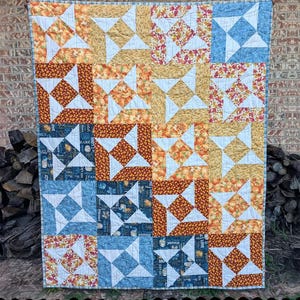 May include: A handmade quilt with a patchwork design. The quilt features squares with various patterns, including pumpkins, floral prints, and blue geometric shapes. The text "Tall Windmills" is at the top, and "Sewl Sister for Villa Rosa Designs" is at the bottom.