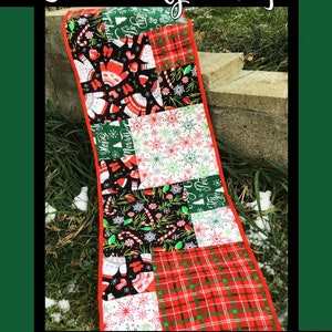 May include: A red and green Christmas table runner with a patchwork design featuring various holiday patterns, including snowflakes, candy canes, and the words "Hope" and "Merry". The table runner is made by Tricia Lynn Maloney for Villa Rosa Designs.