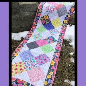 May include: A colorful table runner with a criss-cross pattern. The runner is made of fabric with floral prints and is quilted. The runner is on a background of snow and grass. The text "Criss Cross" is at the top of the image. The text "Tricia Lynn Maloney Orphan Quilt Designs" is at the bottom of the image. The text "For Villa Rosa Designs" is at the bottom of the image.