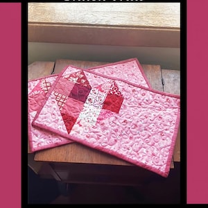May include: Two pink and red quilted snack mats with a heart design. The mats are made by Tricia Lynn Maloney for Orphan Quilt Designs and Villa Rosa Designs.