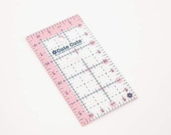 Könnte beinhalten: Ein rechteckiges Quilt-Lineal aus klarem Acryl mit rosa Rändern und schwarzen Rastermarkierungen. Das Lineal trägt den Text Cute Cuts by Lori Holt of Bee in my Bonnet, mit imperialen Maßen in Zoll und kleinen Blumen-Symbolen an den Rändern.