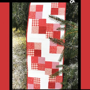 May include: A red and white patchwork quilt table runner with a snowflake design. The quilt is made of various fabrics, including gingham, polka dots, and floral prints. The quilt is called "Snowberry" and is designed by Tricia Lynn Maloney for Villa Rosa Designs.