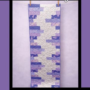 May include: A quilted wall hanging with the text "Heart Strings" in a script font. The quilt features a patchwork design in shades of purple and white, with a textured, quilted surface. The design is by Tricia Lynn Maloney for Villa Rosa Designs.