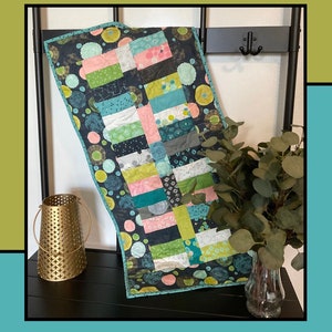 May include: A black and white quilt with a geometric pattern of squares and circles. The quilt is made with a variety of fabrics in shades of green, blue, pink, and white. The quilt is hanging on a black metal rack with a gold metal container and a vase of green leaves.