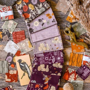 May include: Assortment of patterned fabric pieces and folded bundles in shades of burgundy, brown, and lavender, featuring floral and botanical patterns. A vintage bird illustration and decorative items are arranged on a wooden surface.